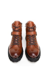 Botas de ante de cuero marrón hasta el tobillo con suelas negras, con ojales de metal, cordones marrones y una cremallera lateral. Textura suave, diseño robusto.