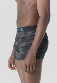 Torso masculino con bóxer en camuflaje negro y gris, con patrones de tiburón azul y cintura negra con letras azules.