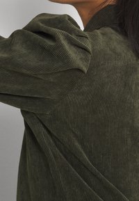 Top in corduroy verde oliva con maniche a sbuffo e texture a righe verticali, con un design drappeggiato e dettagli di cucitura diagonali.