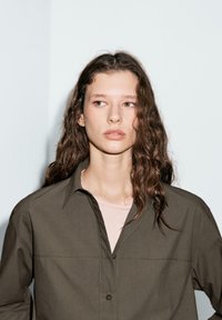 Olive green button-up shirt with a pointed collar, long sleeves, and a relaxed fit. Worn over a light pink top, featuring textured hair.