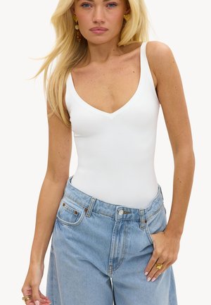 Femme blonde portant des boucles d'oreilles en or, un haut blanc sans manches en V et un jean bleu clair ample taille haute avec des bagues aux deux mains.