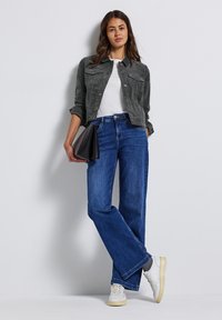 Žena opřená o zeď, oblečená v tmavě zelené bundě, bílé tričko, modré džíny, bílé tenisky a držící černou psaníčko.