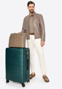 Valigia rigida verde scuro con texture a righe, borsa per laptop strutturata marrone sopra, indossata da un uomo in polo beige e pantaloni chiari.