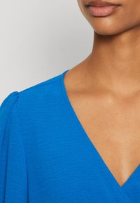 Halenka s výstřihem do V v jasně modré barvě, vyrobená z texturované látky. Na rukávech je detail s balónovými efekty a kolem výstřihu je čistě prošívaný okraj.