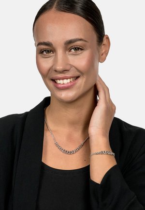 Silberne Kettenhalskette und Armband mit einem verknüpften Design, getragen von einem Modell in einem schwarzen Oberteil, das ihren Glanz vor einem neutralen Hintergrund hervorhebt.