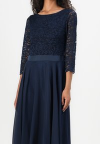 Robe bleu marine avec un corsage en dentelle aux motifs floraux et manches trois-quarts, jupe fluide à la texture lisse, et ceinture en satin.