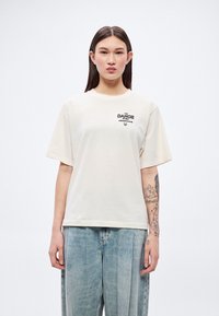 Krémové tričko s krátkym rukávom a okrúhlym výstrihom s čiernym potlačeným nápisom "THE DANCE STUDIO." Kombinované so svetlomodrými voľnými džínsami.