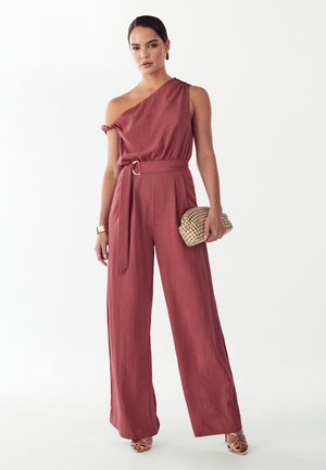 Žena v jednoramennom fialovom kombinéze s opaskom, drží pletenú kabelku, stojí pred jednofarebným bielym pozadím.