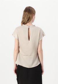 Beige top s krátkým rukávem z lehkého materiálu, se designem na zádech ve tvaru klíčové dírky a knoflíkovým zapínáním, s oblým lemem.