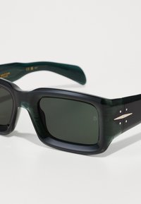 Ochelari de soare cu un cadru negru lucios, lentile de culoare verde închis și accente metalice pe temple. Formă pătrată cu brațe groase.