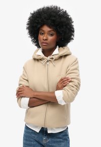 Felpa beige con cappuccio e cordino, zip frontale e tasche laterali, abbinata a una camicia bianca con colletto e jeans di denim. Texture morbida, design casual.