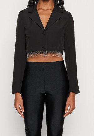 Žena oblečená v černém kratším blejzru s třásněmi z kamínků podél lemu a v černých vysokým pasem kalhotách na jednobarevném pozadí.