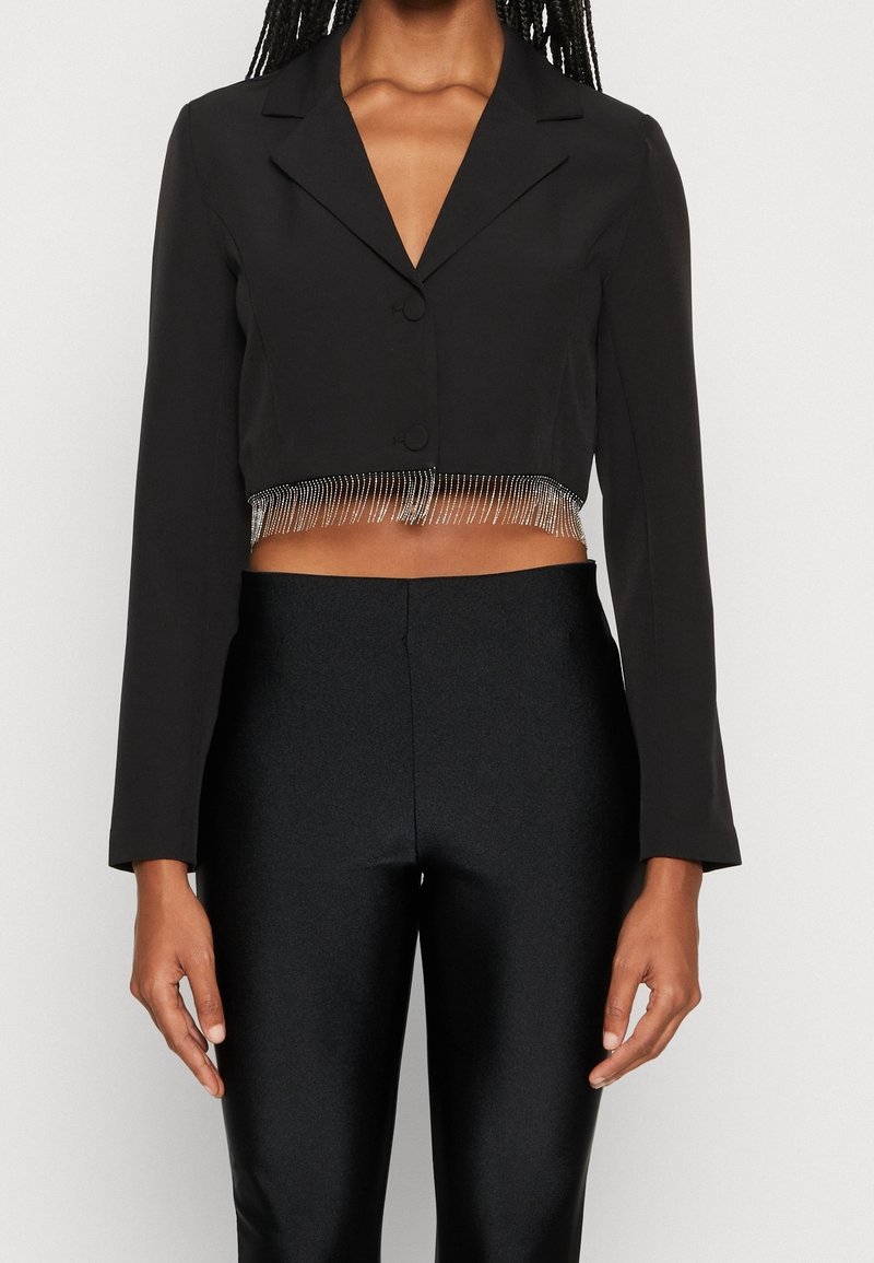 Femme portant un blazer court noir orné d'une frange de strass le long de l'ourlet et un pantalon noir taille haute, sur un fond uni.