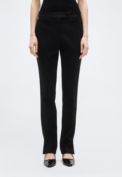 Femme portant un pantalon noir sur mesure et des talons pointus en cuir verni noir, debout devant un fond clair uni.