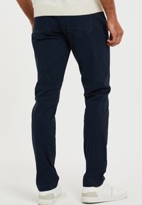 Marineblaue Baumwoll-Chinos mit einem geraden Schnitt, ausgestattet mit Gesäßtaschen und kombiniert mit weißen Sneakers. Glatte Textur und klare Linien.
