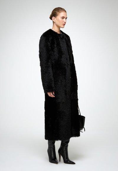 Cappotto lungo in pelliccia sintetica nera con ampio colletto, caratterizzato da una chiusura con bottoni. Abbattutto con stivali neri a punta, al ginocchio.