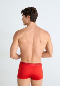 Boxer shorts rojos con una textura suave y una cinturilla elástica que presenta el logo de "Calvin Klein" en negro. Se muestra al modelo de espaldas.