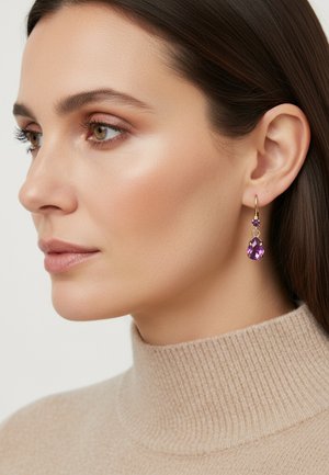 Boucles d'oreilles créoles en or ornées de pierres précieuses violettes en forme de goutte et de rond. Le modèle porte un col roulé beige qui met en valeur les boucles d'oreilles. Texture de peau lisse.