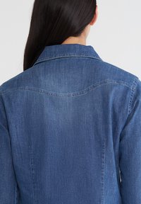 Giacca in denim, blu scuro, con colletto classico e dettaglio a yoke sul retro. Presenta una texture liscia e una vestibilità su misura.