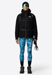 Czarna izolowana kurtka z szarymi paskami, niebieskie legginsy tie-dye i ciemne buty hikingowe. Dodatki to zielone lustrzane okulary przeciwsłoneczne i czarne rękawice.