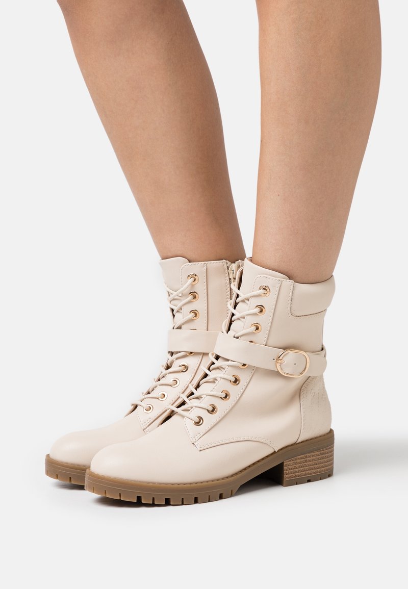Bottes chevilles lacées couleur crème avec bride à boucle, œillets dorés et talon bas épais portées sur jambes nues sur fond blanc.