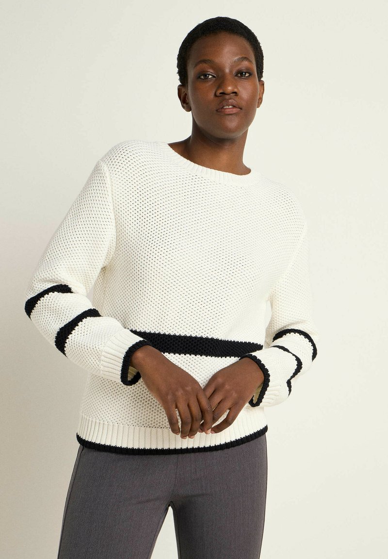 Weißer Strickpullover mit schwarzen Rippakzenten an den Ärmelbündchen und am Saum. Verfügt über einen runden Ausschnitt und eine lockere Passform. Kombiniert mit grauen, eng anliegenden Hosen.