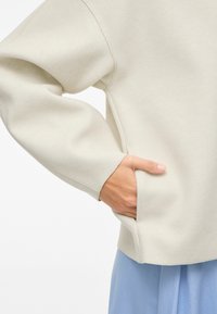 Kreminės spalvos per didelis džemperis, pagamintas iš minkštos medžiagos; turi šoninę kišenę ir atsipalaidavusius rankovės su matomais siuvimo detalėmis.