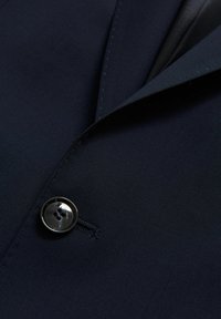 Marinblå kavaj med en slät textur, med en enkel rund knapp med fyra hål och kontrasterande söm på slaget.