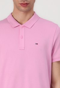 Chemise polo rose en tissu texturé, dotée d'un col classique, d'une patte de boutonnage à deux boutons et d'un petit logo en bleu, blanc et rouge.