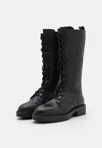 Bottes en cuir noir montantes, avec une texture lisse, présentant un laçage à l'avant et une semelle plate épaisse.