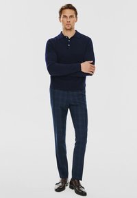 Granatowy sweter dzianinowy z trzykierunkowym zapięciem, zestawiony z granatowymi spodniami w kratkę. Czarne skórzane mokasyny dopełniają stylizację.