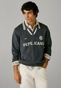 Temno siva pulover s rugby ovratnikom, kremastimi črtami in logotipom "PEPE JEANS". Obut preko kariranega srajčnega oblačila z zavihtimi rokavi.