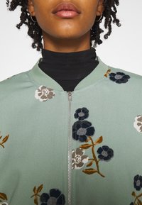 Blumen Bomberjacke aus weichem grünem Stoff, mit Reißverschluss vorne, geripptem Kragen und mehrfarbigen Blumenmustern in Kombination mit braunen, blauen und grauen Akzenten.