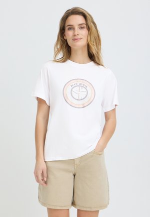 Kvinde med langt blond hår iført en hvid grafisk T-shirt og beige shorts, stående med den ene hånd i lommen mod en ensfarvet baggrund.