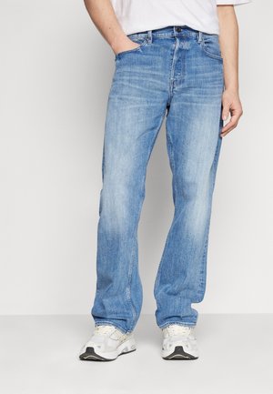 Personne portant un jean droit bleu clair et des baskets blanches, une main dans la poche et une chemise blanche partiellement visible.