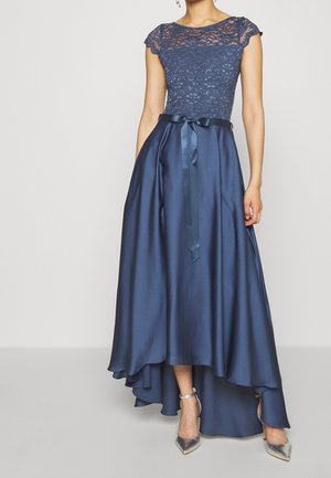 Femme portant une robe bleu marine avec un haut en dentelle, une jupe satinée asymétrique et un ruban noué à la taille, associée à des talons pointus argentés.