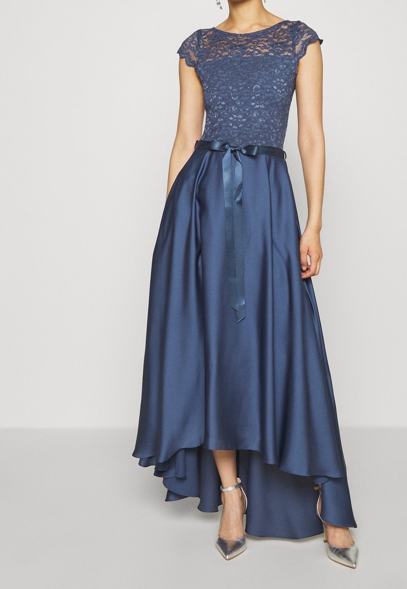 Femme portant une robe bleu marine avec un haut en dentelle, une jupe satinée asymétrique et un ruban noué à la taille, associée à des talons pointus argentés.