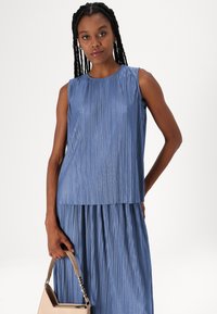 Rochie fără mâneci, plisată, de culoare albastr�ă, cu un design stratificat, având o textură netedă. Modelul ține o geantă bej cu un accent din lanț.