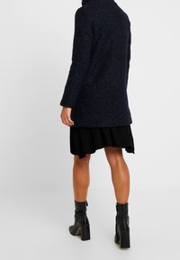 Cappotto in misto lana tessuto navy con colletto alto, silhouette aderente, maniche lunghe e spacco laterale; abbinato a una gonna nera e stivaletti.