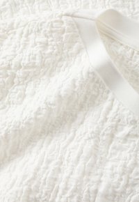 Valkoinen teksturoitu kangas, jossa on aaltoileva kuvio, ja siinä on sileä valkoinen nauha. Pinta on pehmeää, koholla olevaa yksityiskohtaa kauttaaltaan.