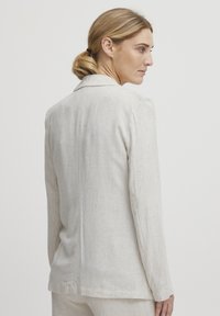 Lyhyt beige pellavablazeri, jossa on klassinen kaulus, pitkät hihat ja sileä rakenne, jossa on hienovarainen kudontakuvio.