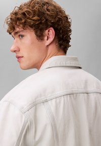 Νέος άνδρας με σγουρά κόκκινα μαλλιά, ντυμένος με ανοιχτόχρωμο τζιν μπουφάν, στραμμένος ελαφρώς στο πλάι μπροστά από ένα απλό γκρι φόντο.