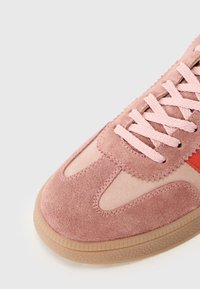 Pink suede sneaker with light pink laces, a smooth light beige panel, and a small orange accent. The sole is textured and brown.