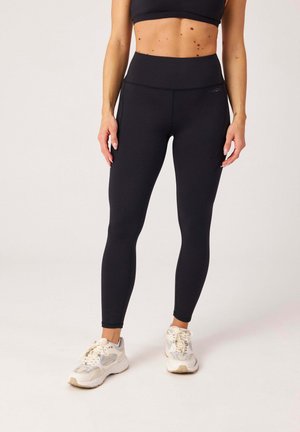 Kobieta ubrana w czarne legginsy z wysokim stanem i białe trampki, stoi z rękami swobodnie opuszczonymi wzdłuż tułowia na jednolitym tle.