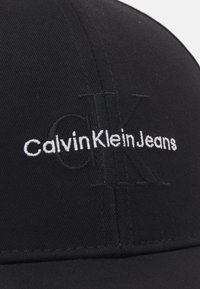 Svart baseballcap laget av bomullsblanding, med forhøyet brodert logo "CalvinKleinJeans" i hvitt. Jevn tekstur med avrundet krone.