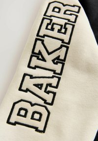 Tejido crema con grandes letras bordadas en negro que dicen "BAKER". El material parece suave y tiene una superficie ligeramente texturizada.