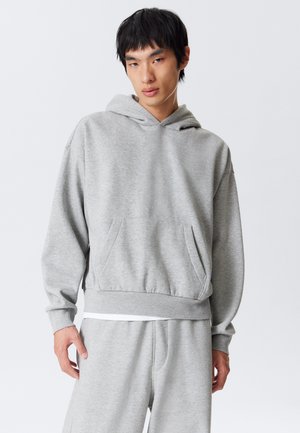 Junger Mann in einem hellgrauen Hoodie und passender Jogginghose, der vor einem schlichten weißen Hintergrund steht.