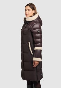 Giacca imbottita marrone con colletto in pelliccia sintetica beige, tasche con cerniera e dettagli a righe sulle maniche; presenta un design lungo e imbottito per il massimo calore.