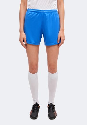 Personne portant un short de sport bleu vif, des chaussettes blanches hautes jusqu'aux genoux et des crampons de football noirs, debout devant un fond uni.