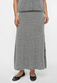Vrouw in een grijze geribbelde gebreide lange rok met zijknopen, grijze top en zwarte ballerina's, staand tegen een witte achtergrond.